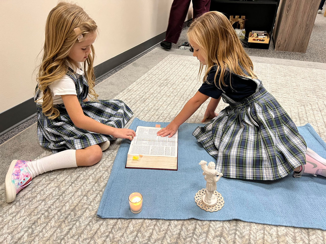 Two young girls reading a religious book on a blue rug.