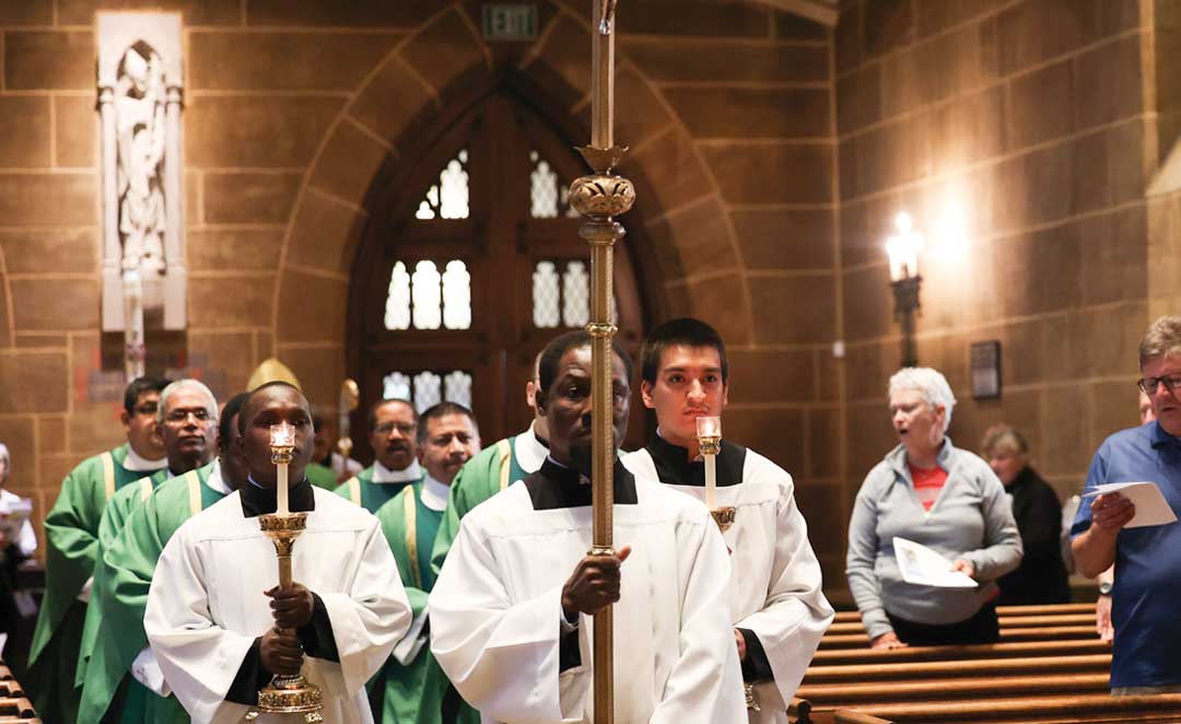 Individuals walking by church pews holding candles with the person in front holding a gold staff.
