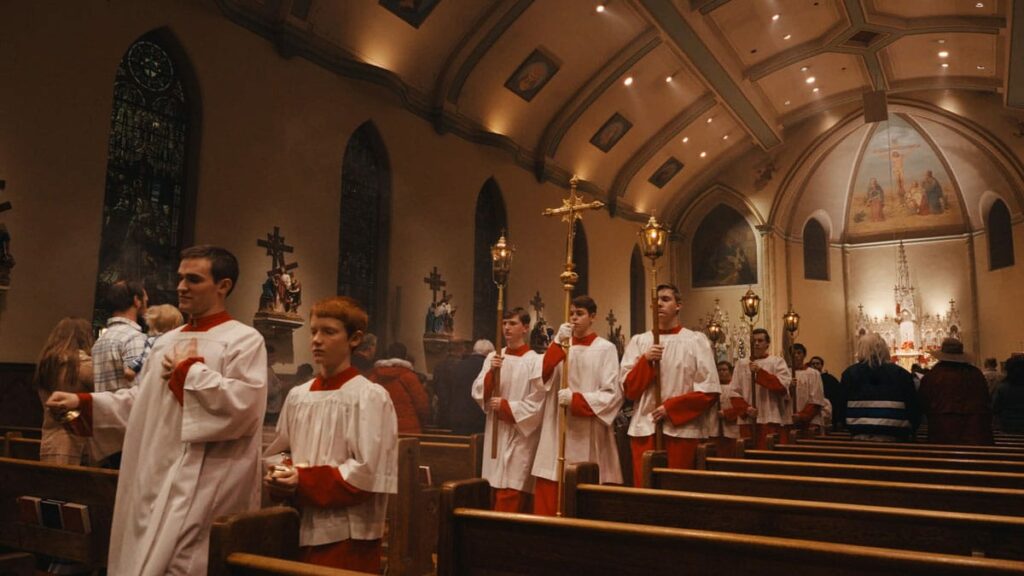 Young men wearing white and red walking down between church pews holding gold staffs and various other items.